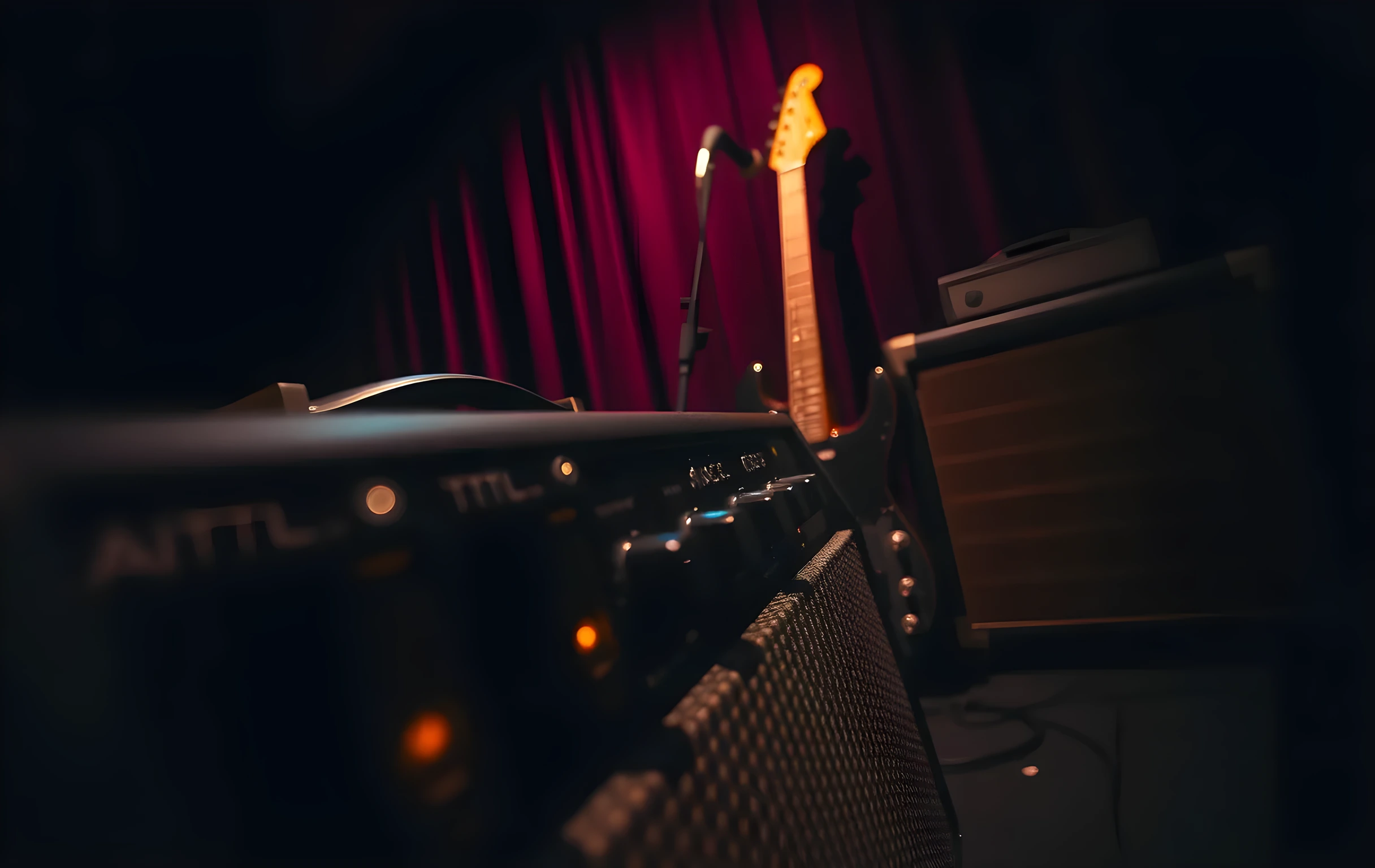 Close detail view of a tube amplifier's control panel with dials and a speaker grille in sharp focus, warm tungsten light reflecting off the metal knobs, the blurred background revealing the edge of a small venue stage where a microphone stand and the silhouette of a bass guitar lean against a dim curtain, moody low-key lighting, no people visible, textures of brushed metal and woven speaker cloth prominent